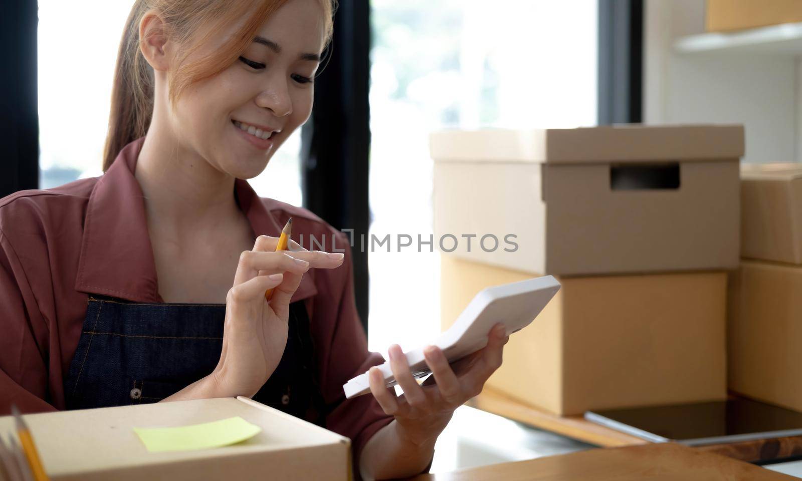 Close up woman sitting in the office full of packages in the background emplaning on a laptop and using a calculator, for online business, SME, finance, e-commerce and delivery concept. by wichayada