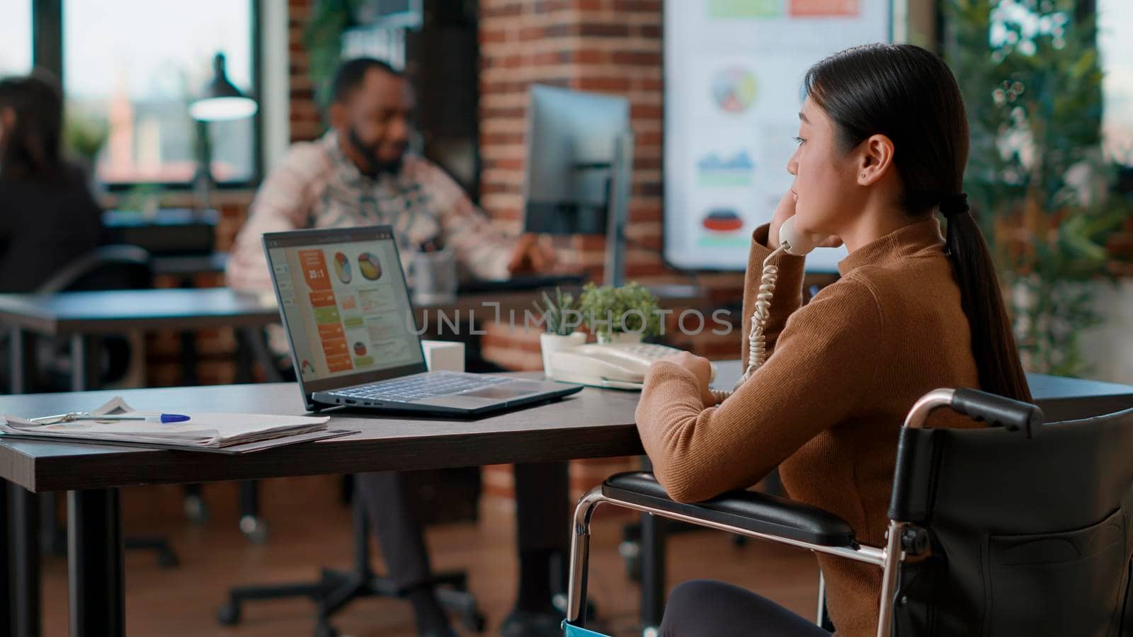 Asian woman in wheelchair answering landline phone and working on business strategy, having remote conversation at work. Employee using office telephone to discuss financial statistics.