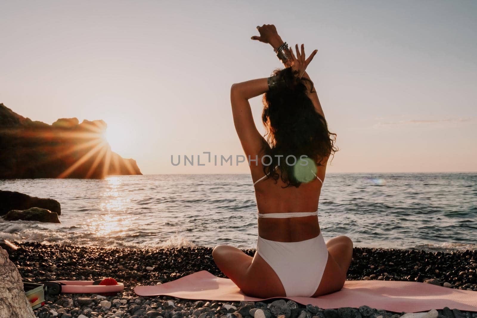Young woman in swimsuit with long hair practicing stretching outdoors on yoga mat by the sea on a sunny day. Women's yoga fitness pilates routine. Healthy lifestyle, harmony and meditation concept.