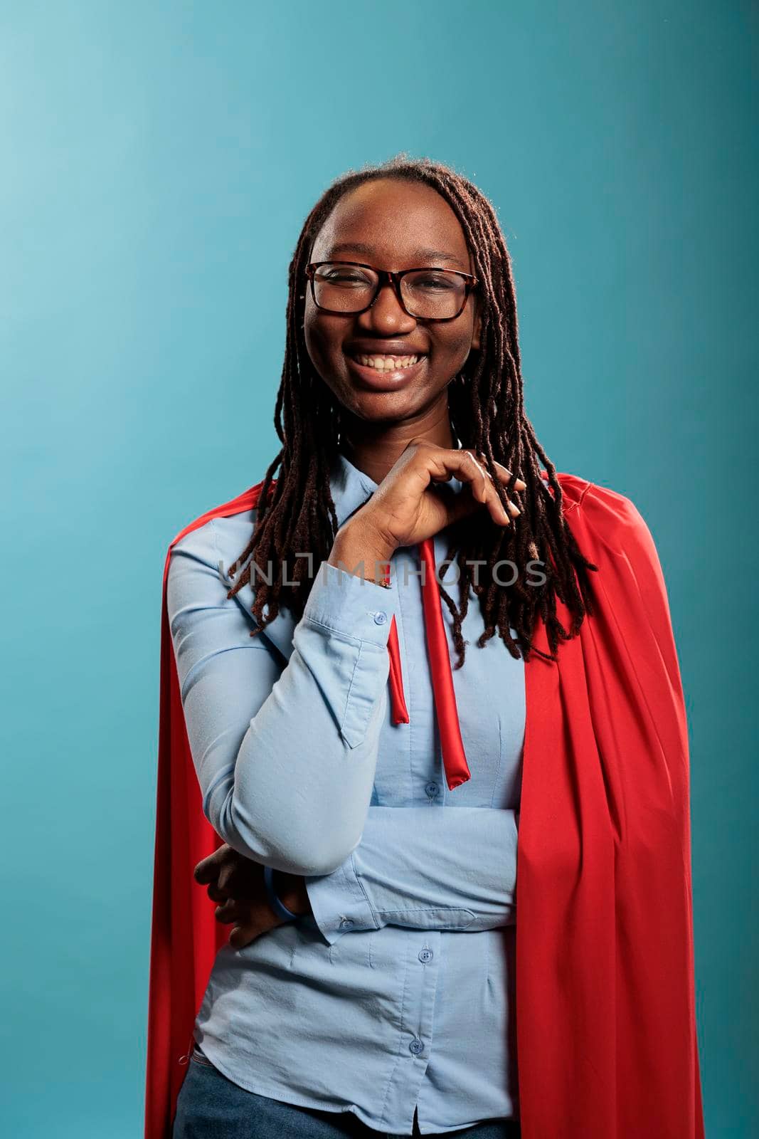 Happy positive smiling justice defender superhero woman wearing hero mighty cape while smiling at camera. Brave and strong person wearing red cloak while standing on blue background. Studio shot.