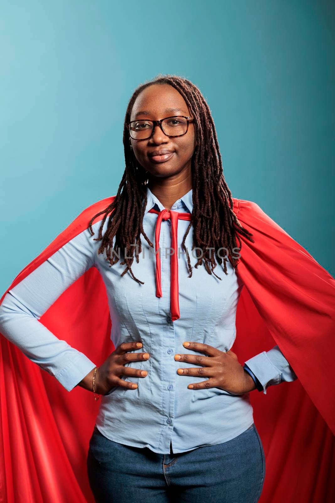 Joyful happy smiling young adult person posing as justice defender on blue background. Proud superhero woman with superpower abilities wearing mighty hero cloak while smiling confident at camera