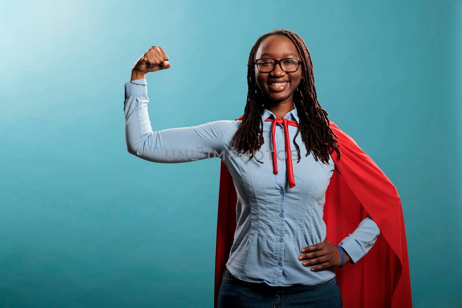 Arms muscle flexing brave superhero woman posing strong and tough for camera on blue background. Justice defender wearing mighty hero red cape while expressing empowerement. Studio shot.