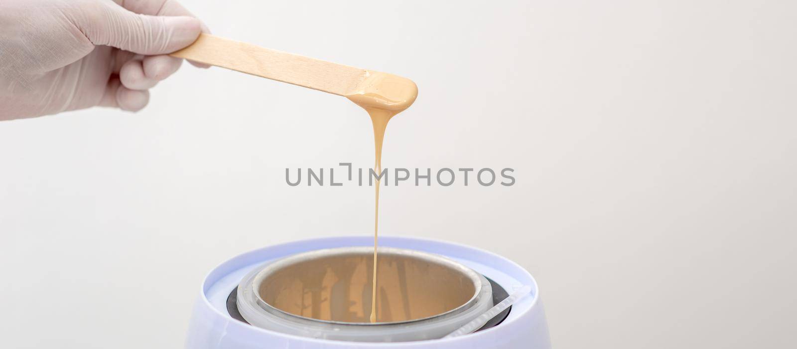 Wax heater with hot yellow wax on wooden spatula in human hand. Hot wax for depilation. Salon equipment