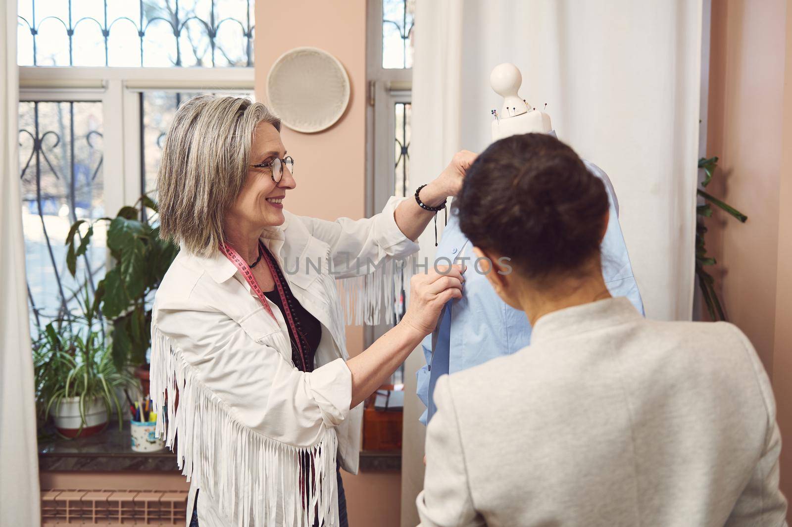 Beautiful mature European female fashion designer and her assistant trying on a new shirt on a mannequin, preparing her new collection of classic clothes for an exhibition in a fashion design studio