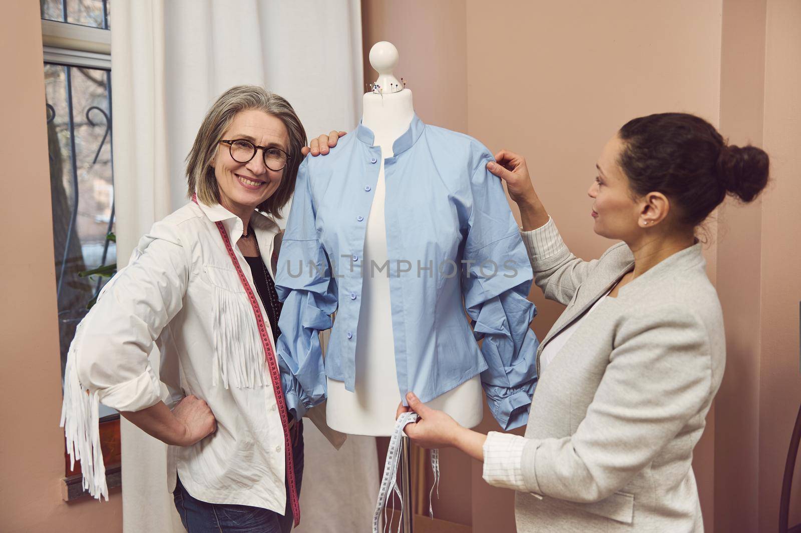 Senior fashion designer smiles posing with a dressmaker model wearing a blue shirt from new collection while her colleague tailor takes measurements and works on improvement details of new garment