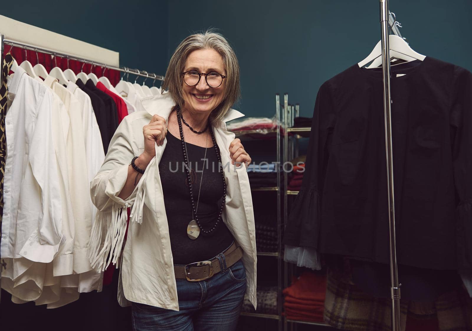 Portrait of smiling successful mature European woman seamstress, dressmaker, tailor posing in own fashion daesign atelier. Fashion designer between hangers with clothing garment in tailoring workshop