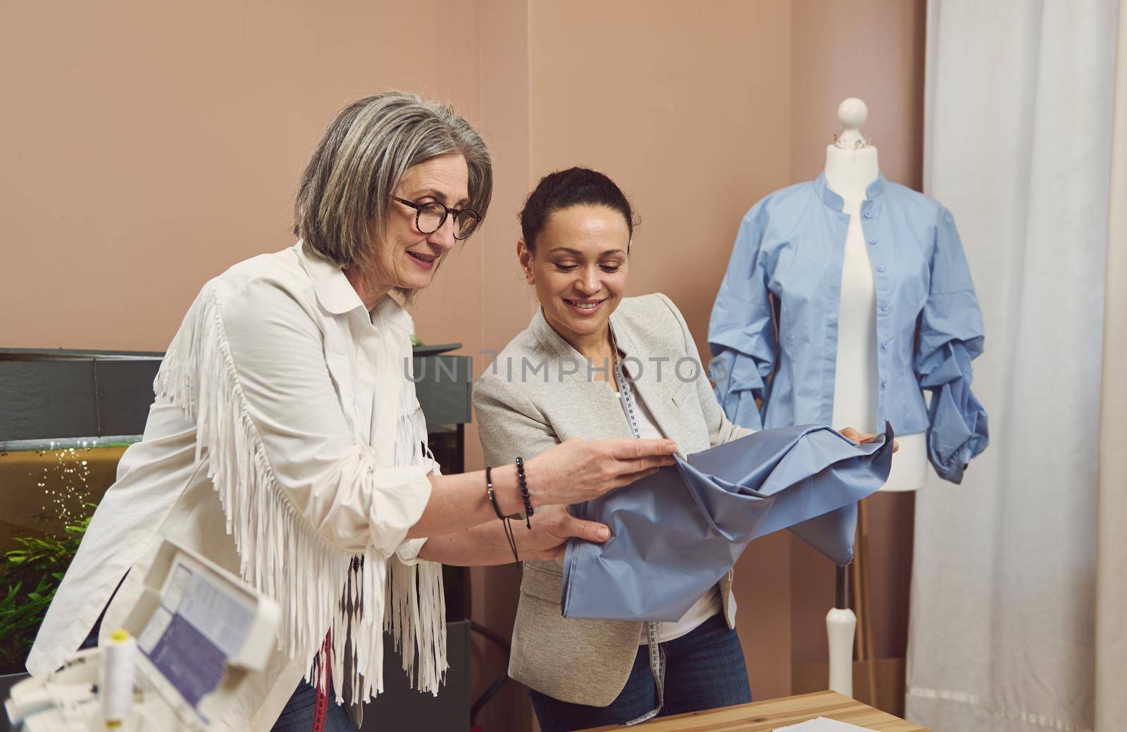 Two seamstresses at work. Skilled fashion designers tailors dressmakers looking at fabric, discussing quality of textile. Colleagues at modern sewing atelier improving production process together