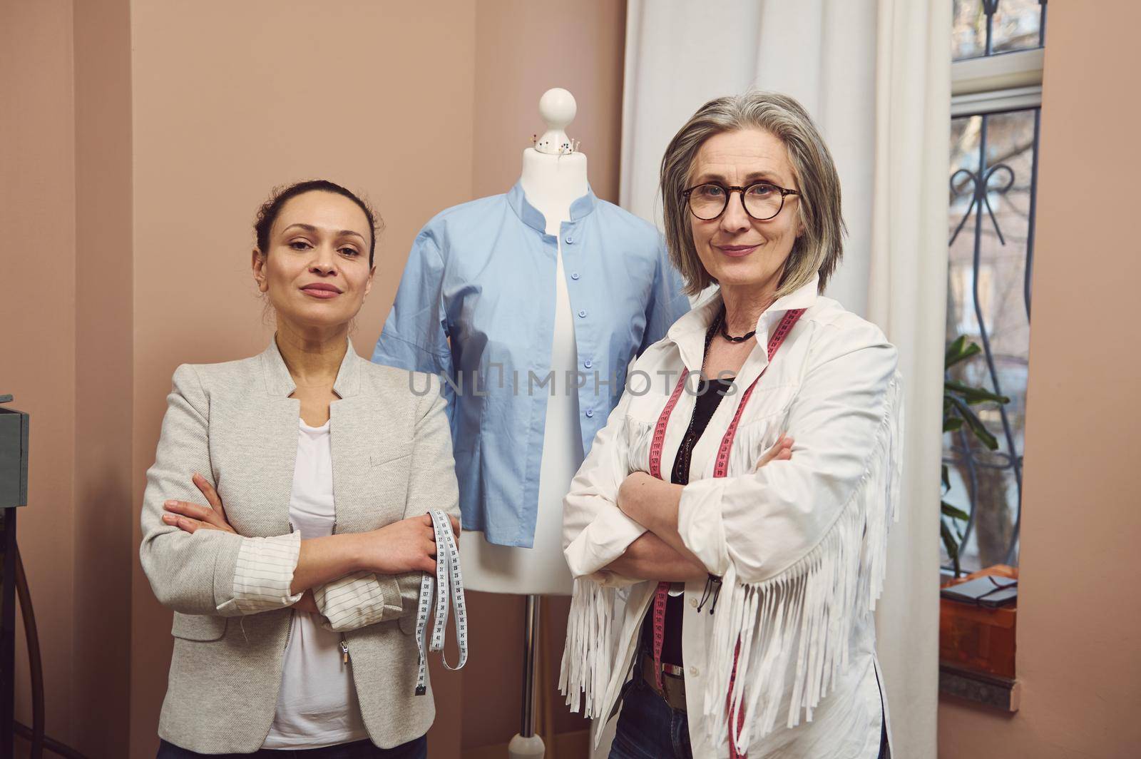 Skilled inspired team of two women seamstresses of different ages and nationalities posing with crossed arms with a mannequin dummy wearing a blue shirt from new collection in a tailoring workshop