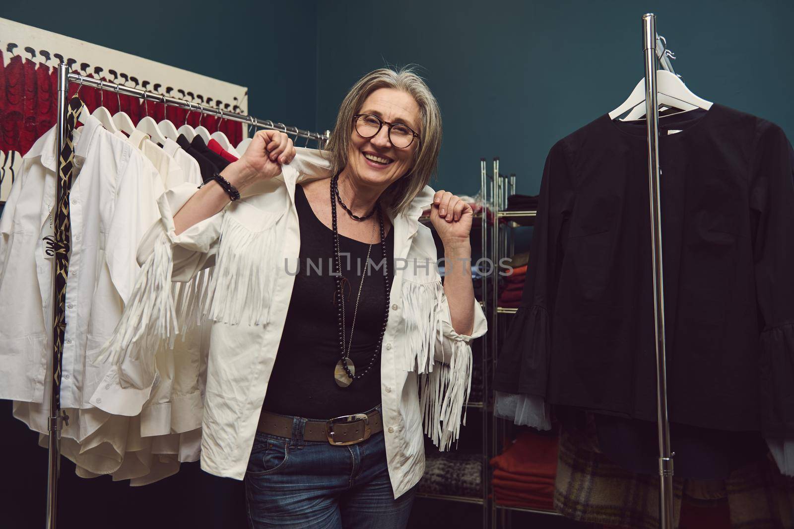 Portrait of happy smiling mature Caucasian woman fashion designer seamstress or tailor posing between hangers with clothes of her new handmade collection in small business fashion or styling workshop.