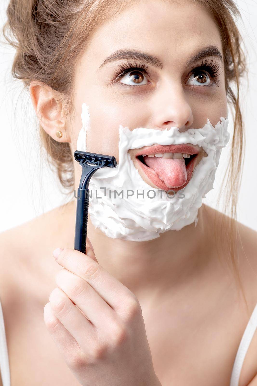 Beautiful young caucasian woman shaving her face by razor on white background. Pretty woman with shaving foam on her face