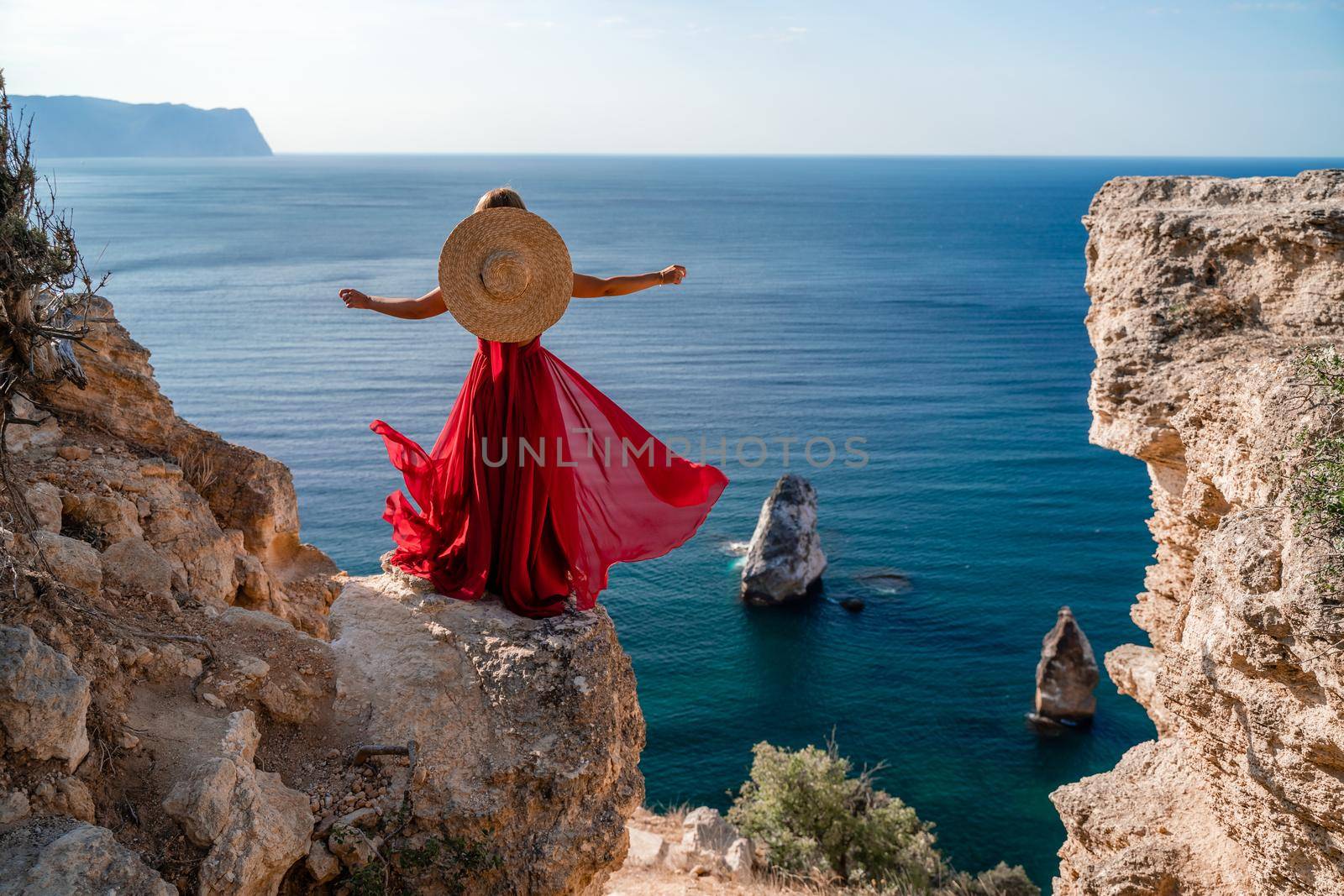 A woman in a flying red dress fluttering in the wind and a straw hat against the backdrop of the sea