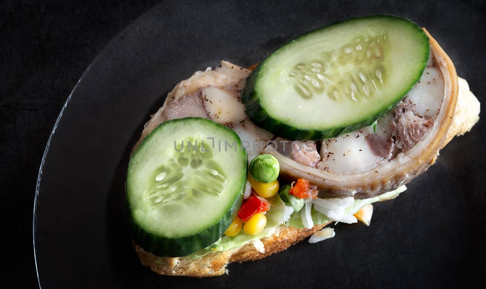 On a glass plate is a sandwich with a meat snack and vegetables. Top view, dark background, copy space