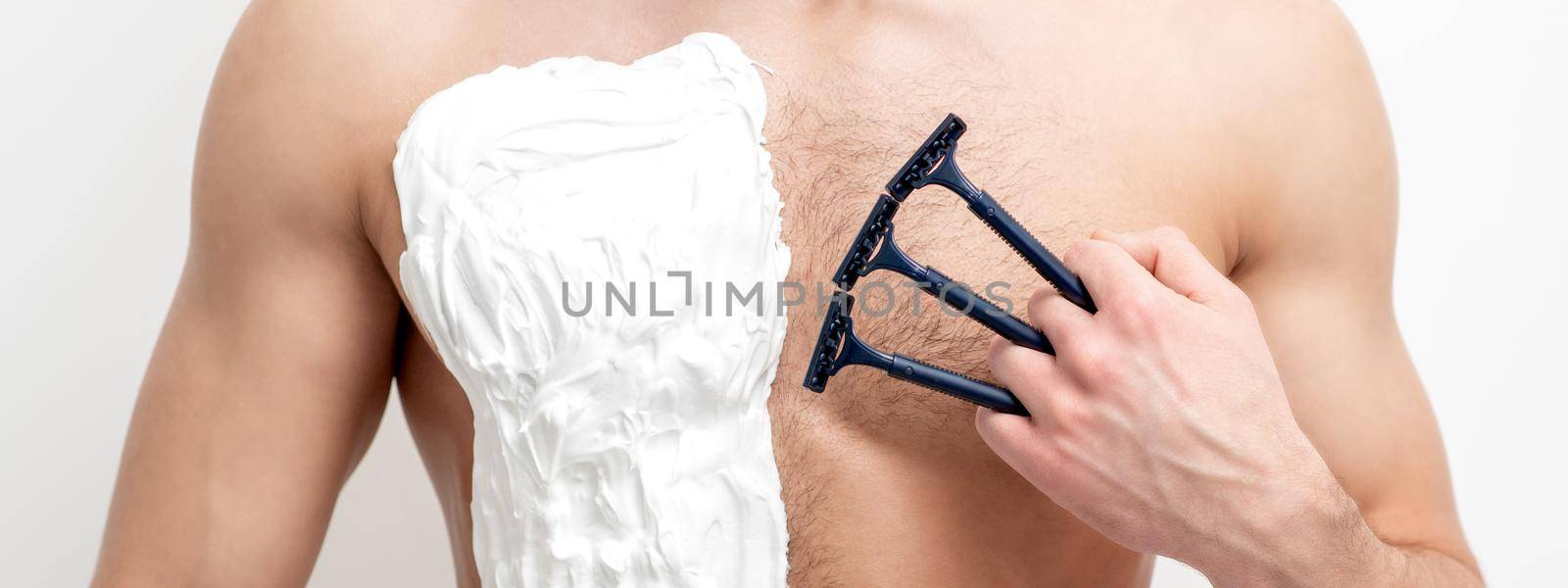 Young caucasian man with beard holds razor shaves his chest with white shaving foam on white background. Man shaving his torso