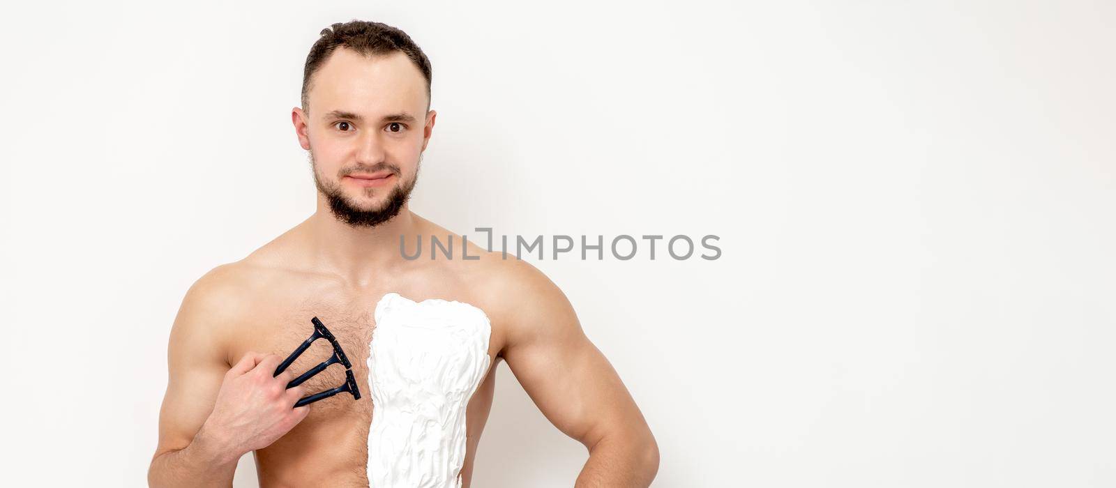 Young caucasian man with beard holds razor shaves his chest with white shaving foam on white background. Man shaving his torso