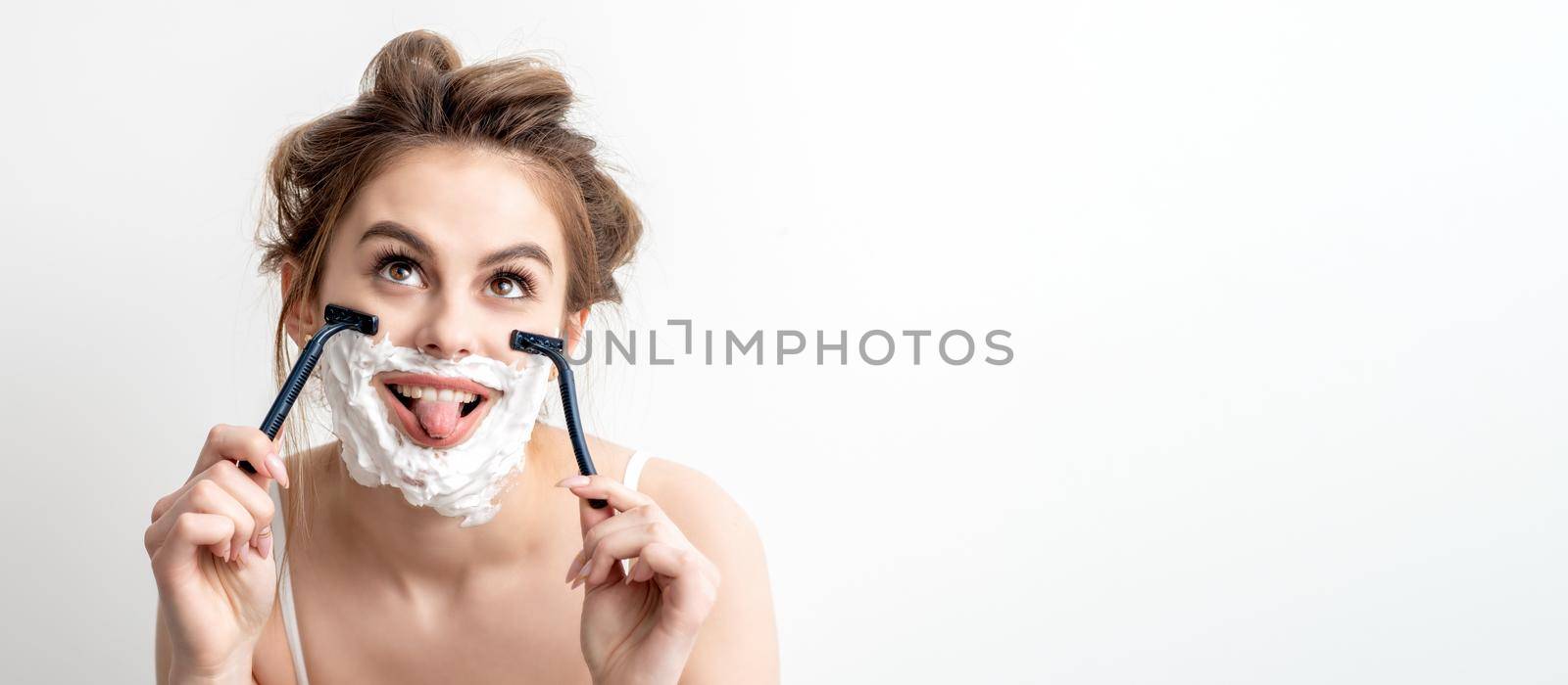 Beautiful young caucasian woman shaving her face by two razors with tongue out on white background.