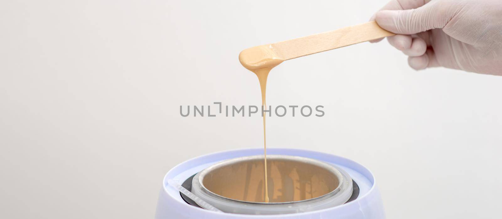 Wax heater with hot yellow wax on wooden spatula in human hand. Hot wax for depilation. Salon equipment