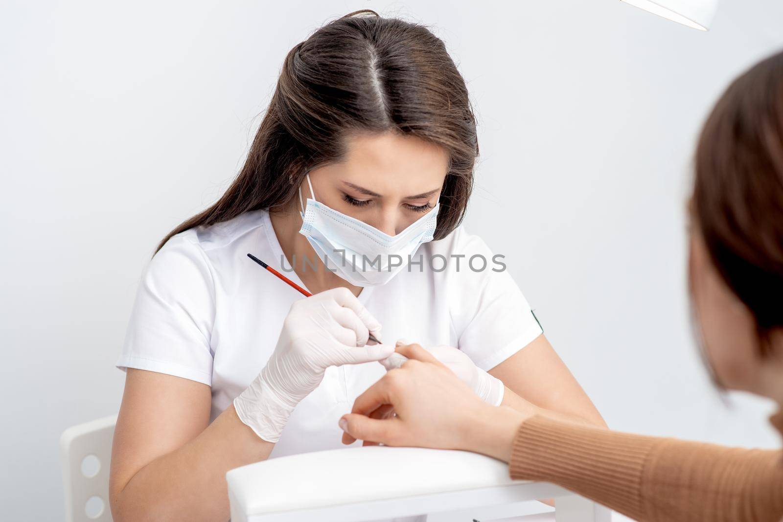 Manicure master with protective mask applying transparent nail polish on female nails in beauty salon