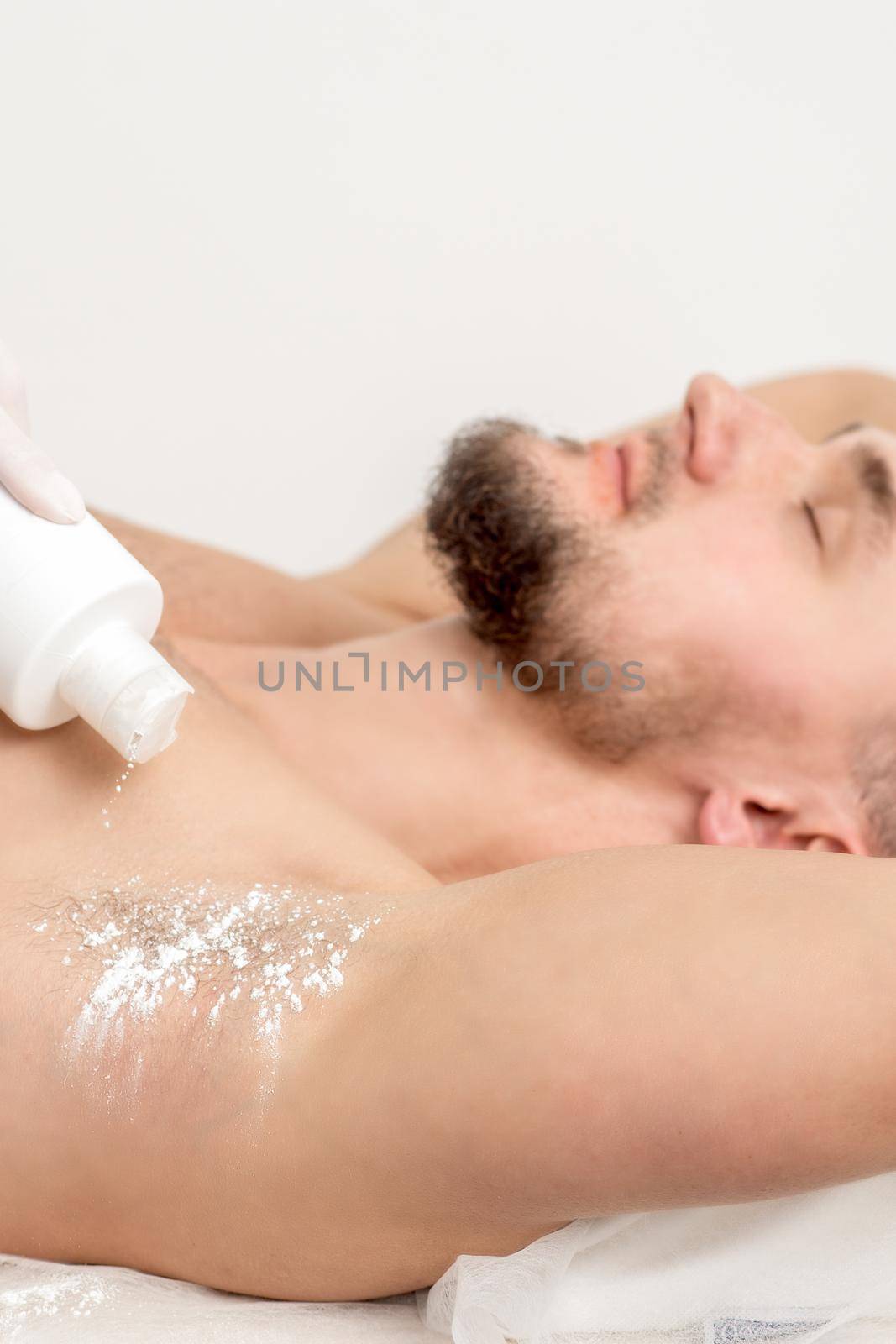 Hand of master depilation pouring talcum powder on armpit of young man before depilation procedure.