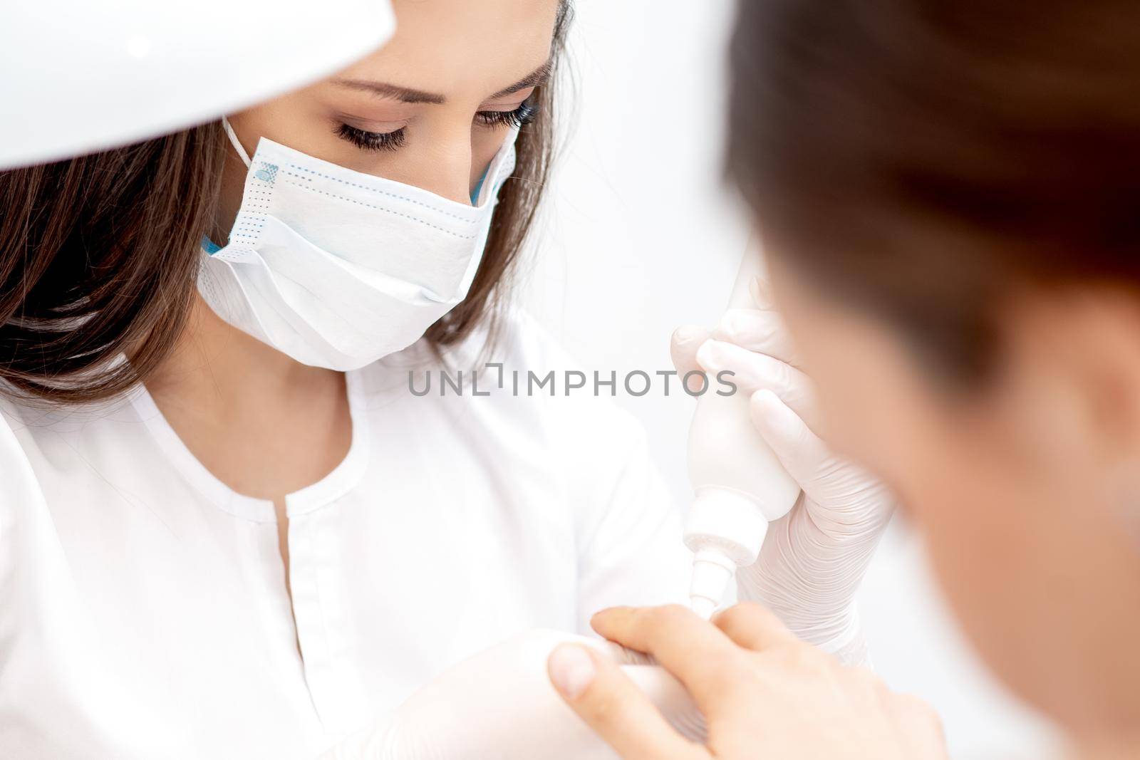 Professional manicurist with protective mask pouring oil on nails manicure of woman in beauty salon.