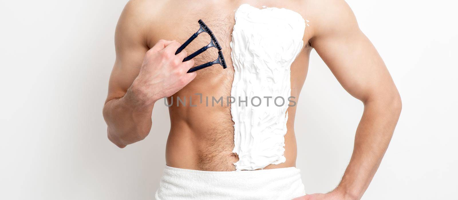 Young caucasian man with beard holds razor shaves his chest with white shaving foam on white background. Man shaving his torso