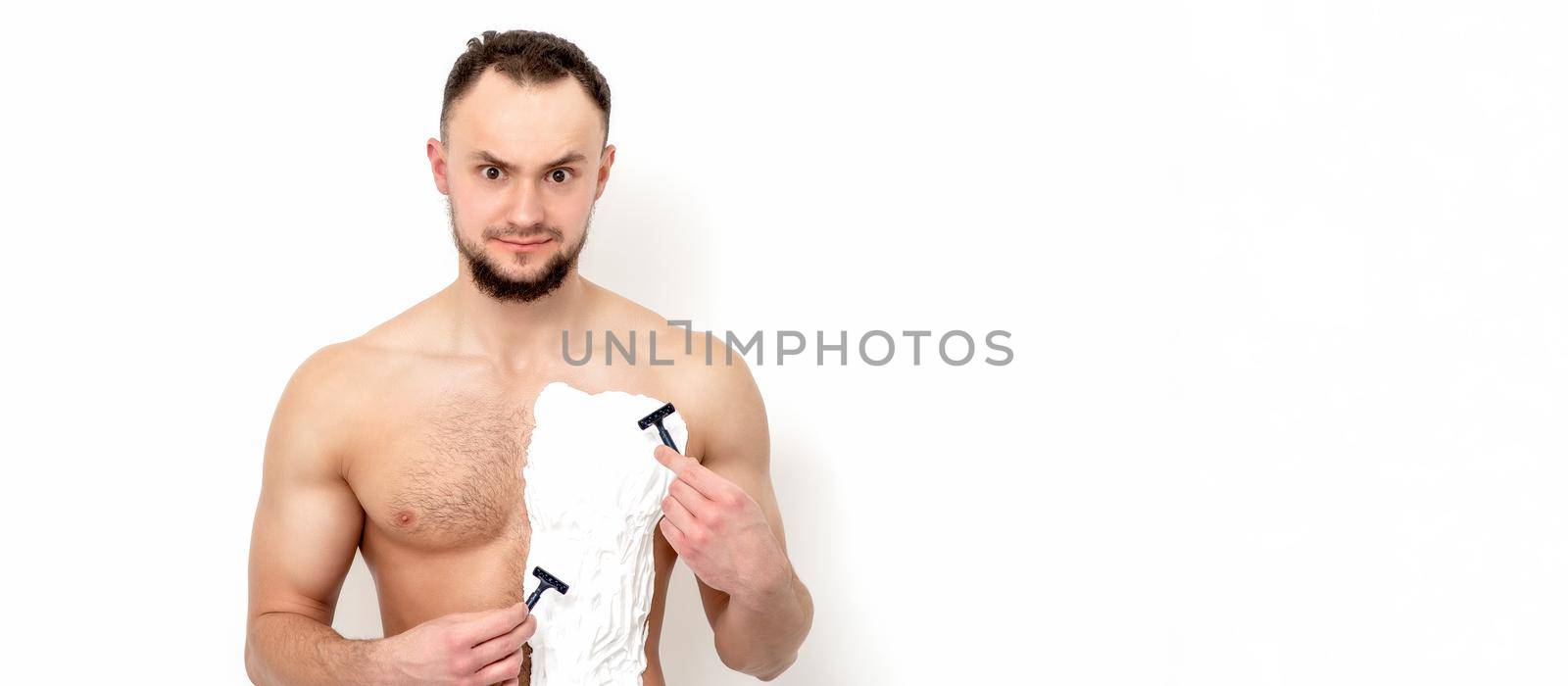 Young caucasian man with beard holds razor shaves his chest with white shaving foam on white background. Man shaving his torso