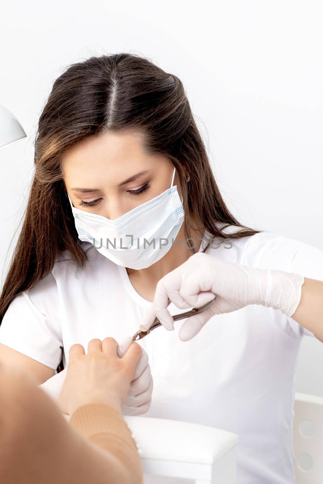 Manicurist in protective mask using manicure cutter to remove cuticle of female nails in manicure salon