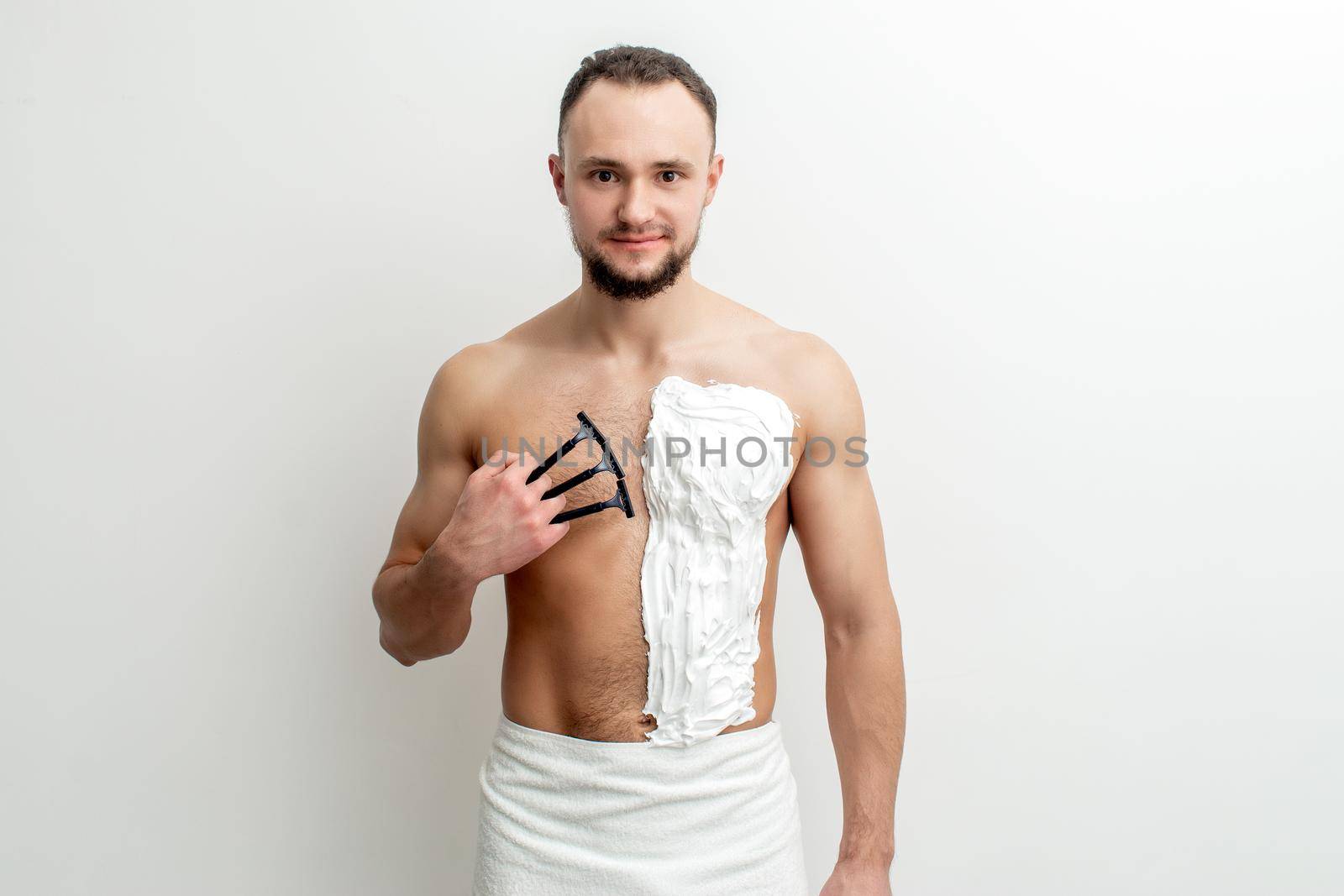 Young caucasian man with beard holds razor shaves his chest with white shaving foam on white background. Man shaving his torso