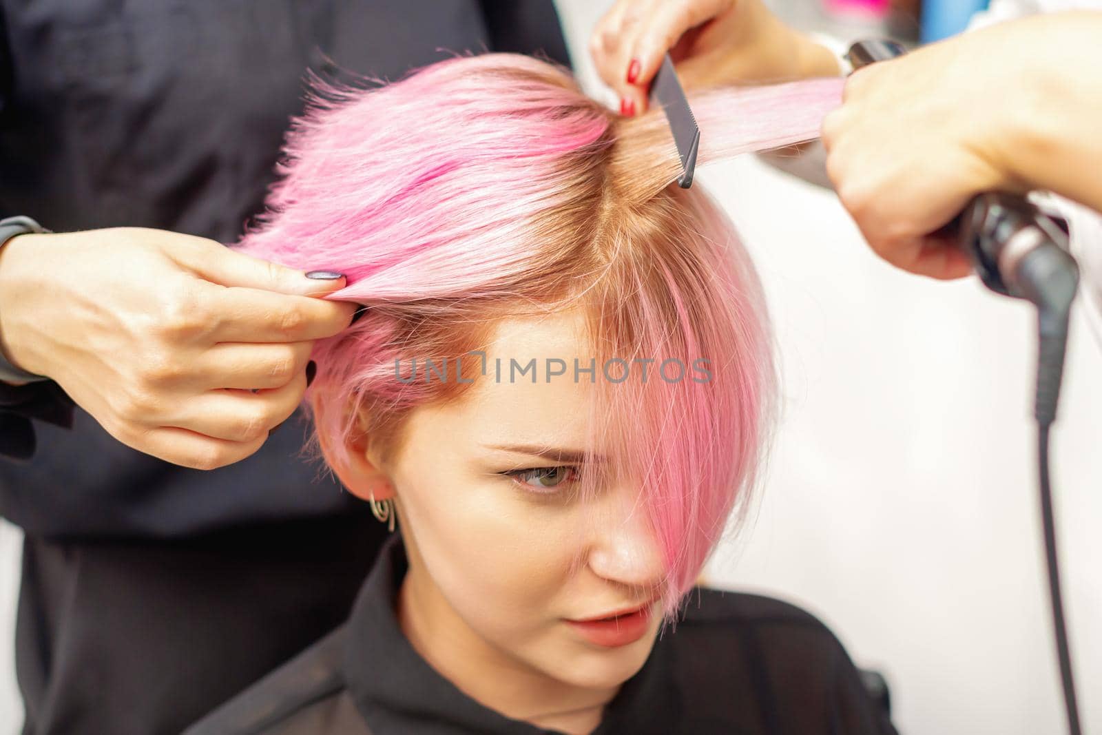 Hairdresser straights pink hair of young woman by hair iron in hairdress salon