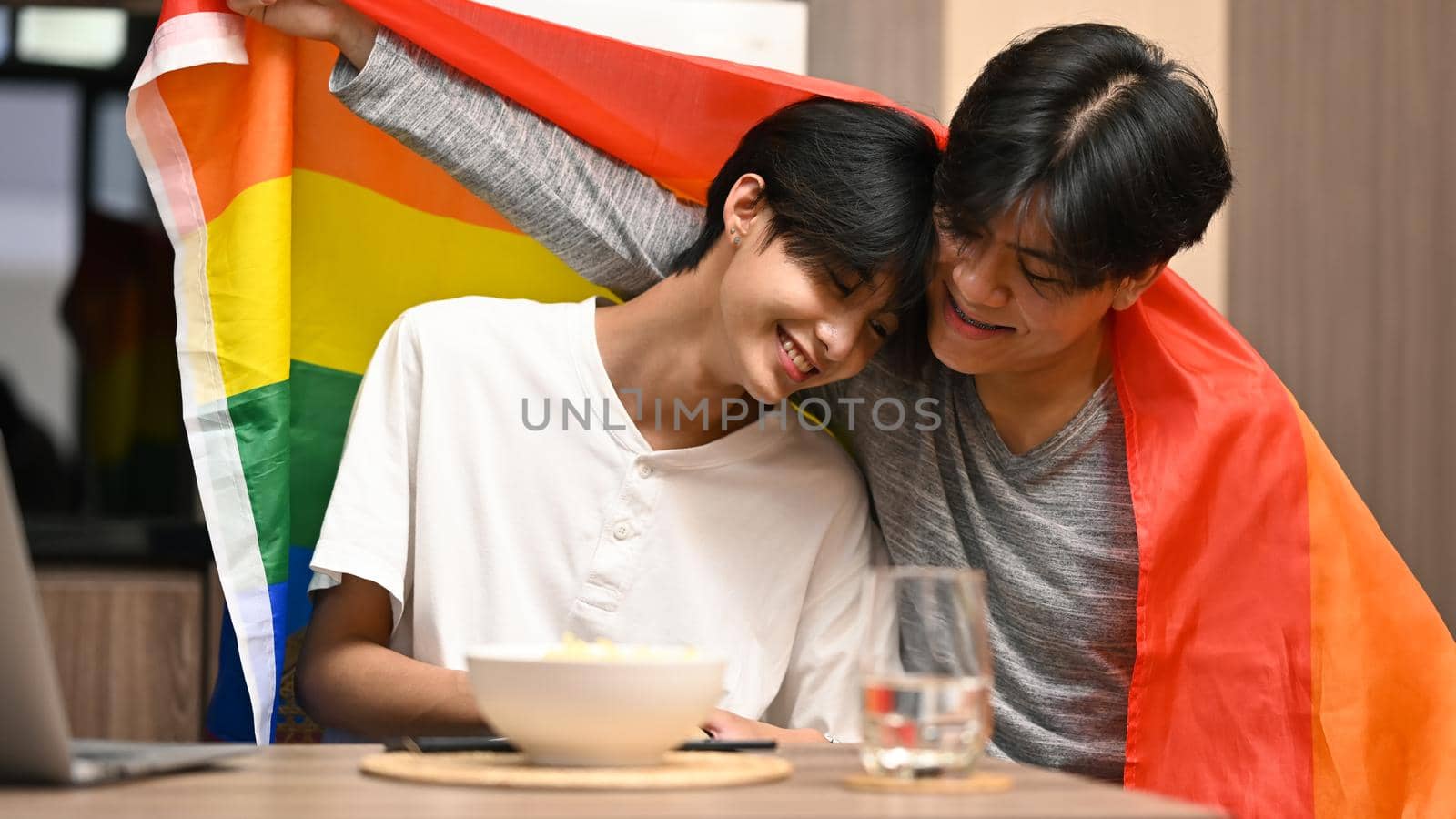 Happy young homosexual couple embracing under LGBTQ pride flag. Concept of sexual freedom and equal rights for LGBT community.