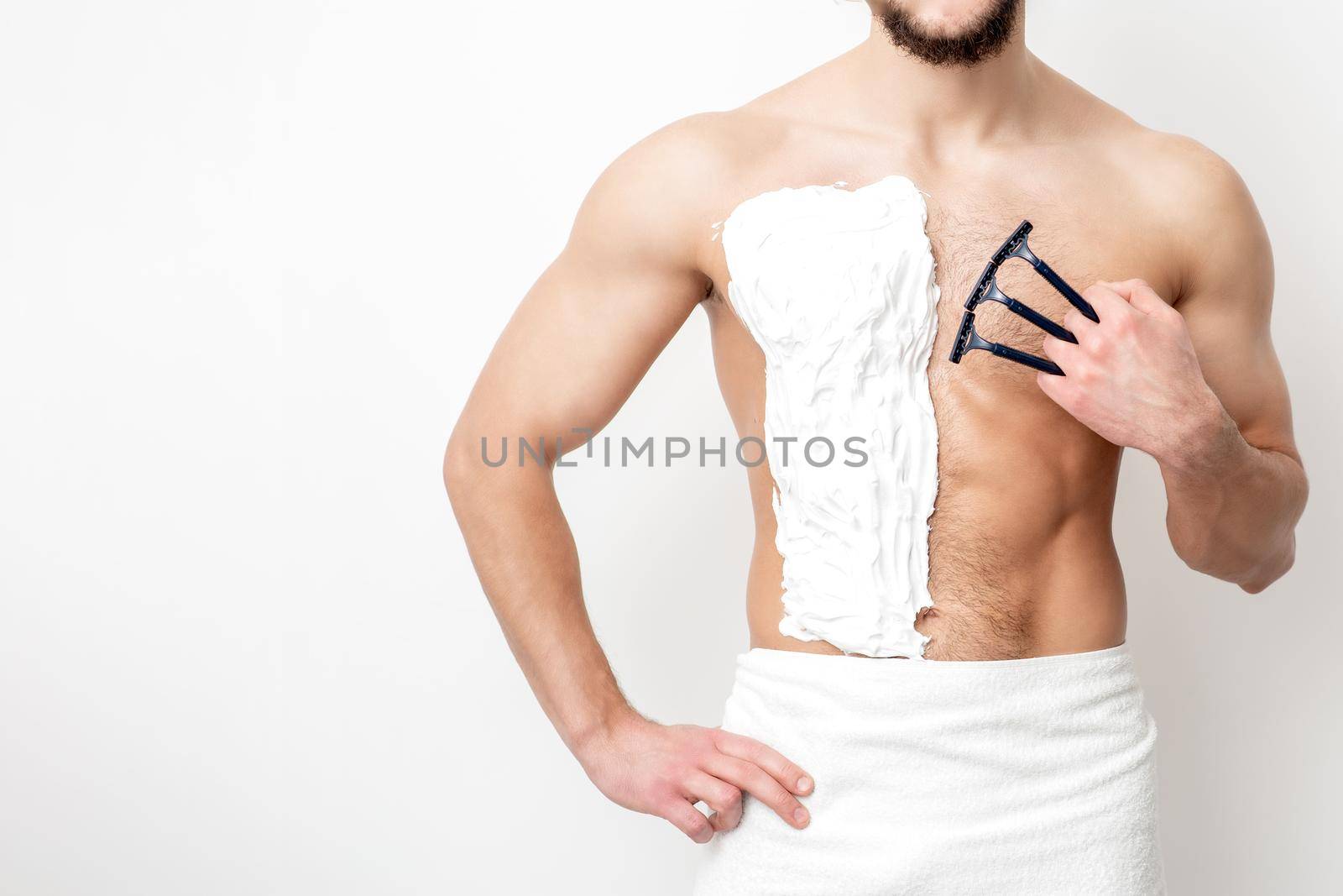 Young caucasian man with beard holds razor shaves his chest with white shaving foam on white background. Man shaving his torso