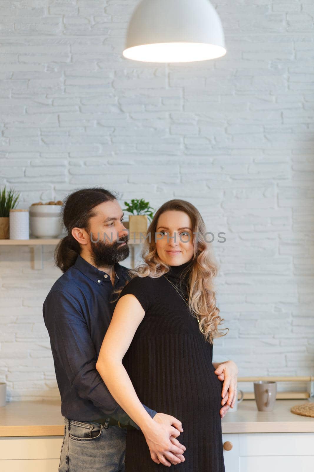 Man embracing pregnant partner in kitchen. Adult family pregnancy concept. Future parents in home outfit embrace standing in the kitchen, looking at each other, kissing. Healthy Lifestyle.