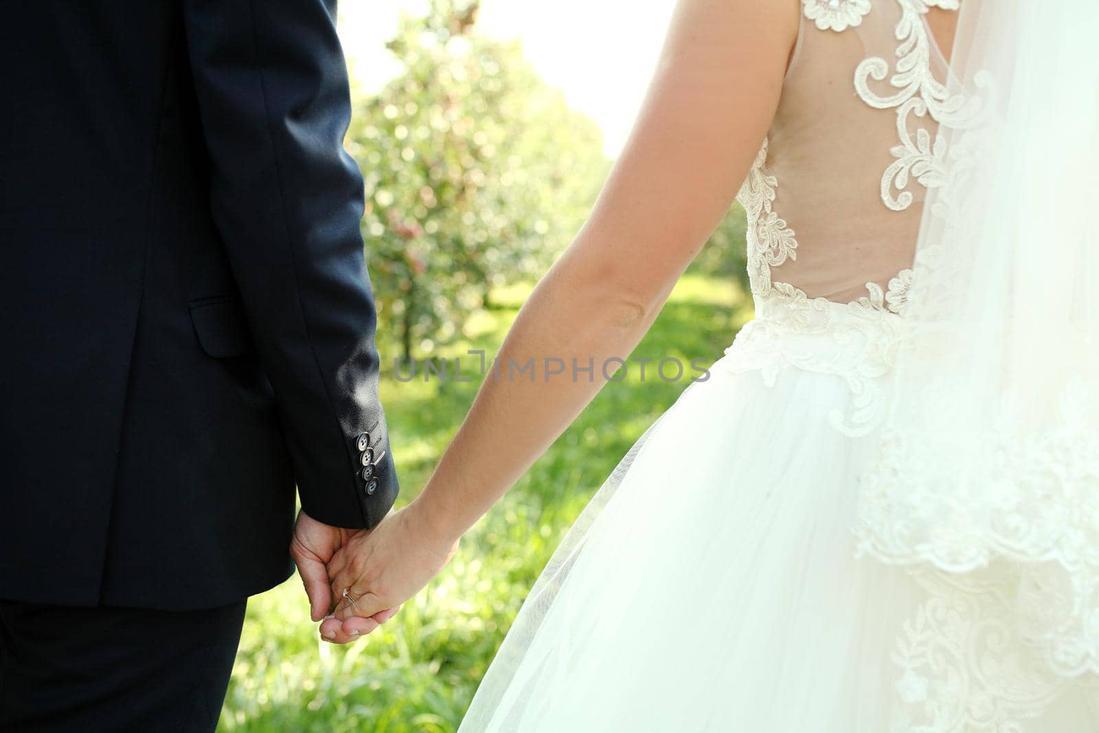 Back view of bride in white dress and groom in suit holding each others hands outdoors