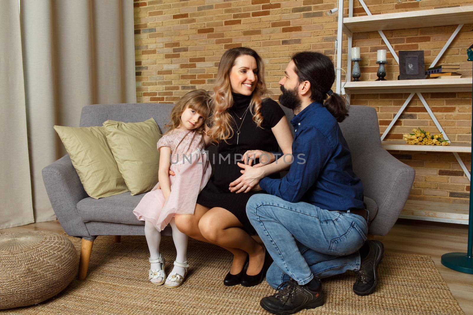 Happy young father, mother and daughter sit on wicker sofa at home. The image of a happy family expecting the second child, studio