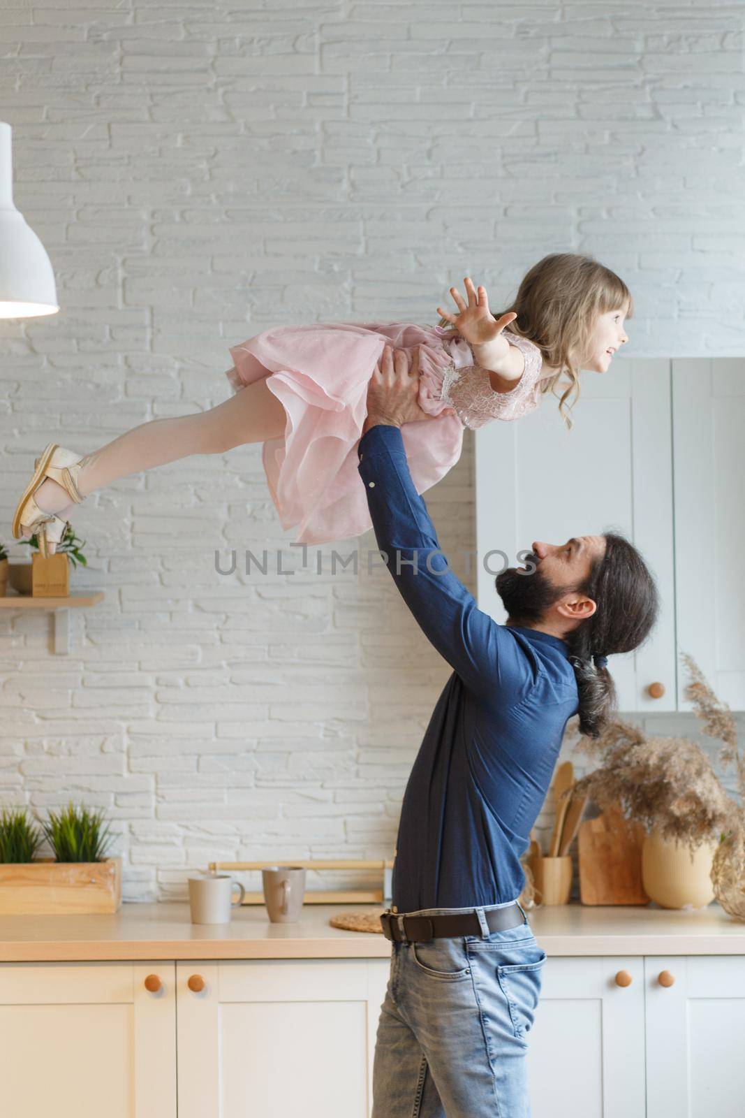Happy young man lifting small preschool child daughter in air standing in modern studio living room. Overjoyed kid girl having fun with handsome father, flying on hands at home.