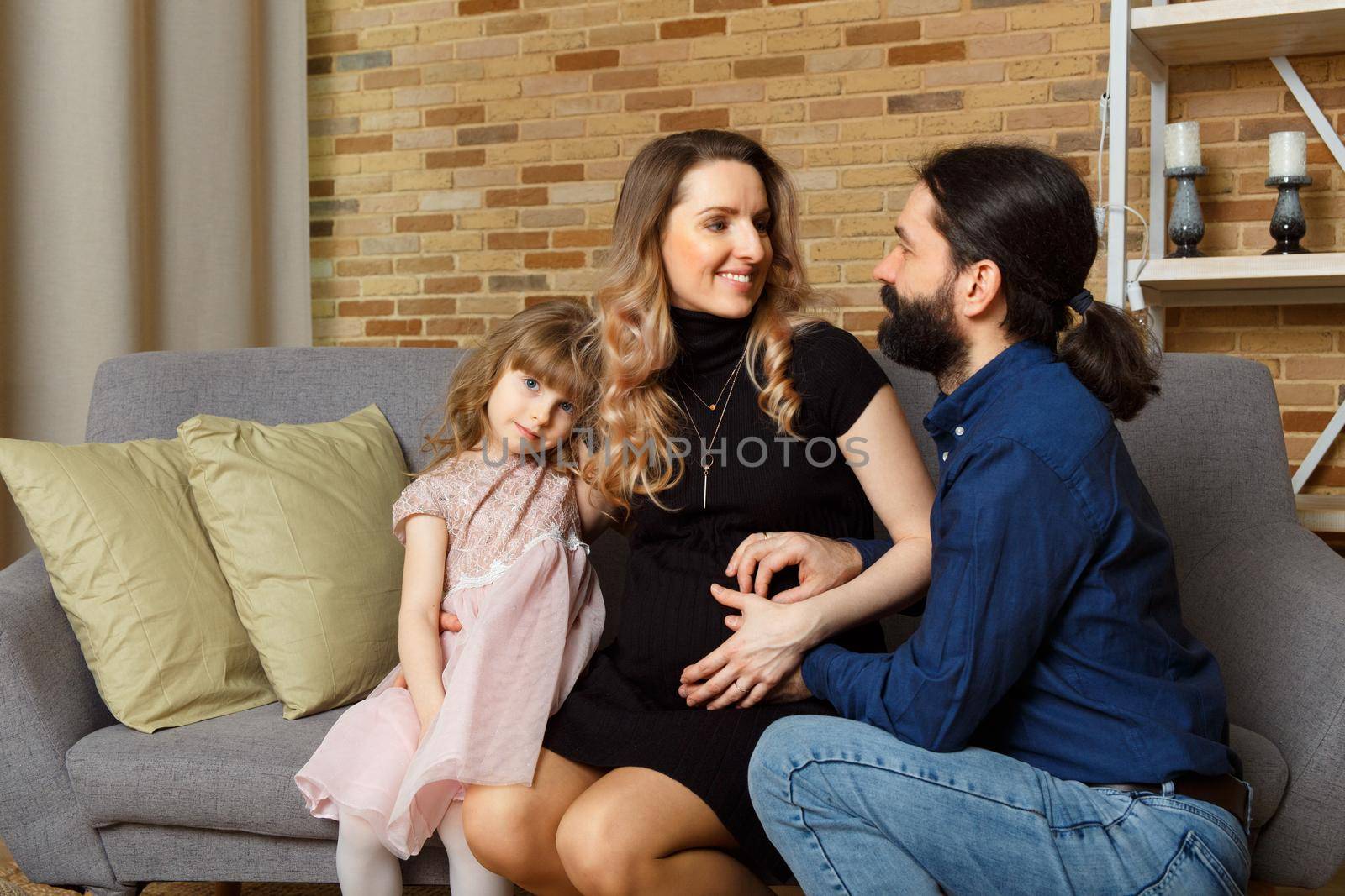 Happy young father, mother and daughter sit on wicker sofa at home. The image of a happy family expecting the second child, studio