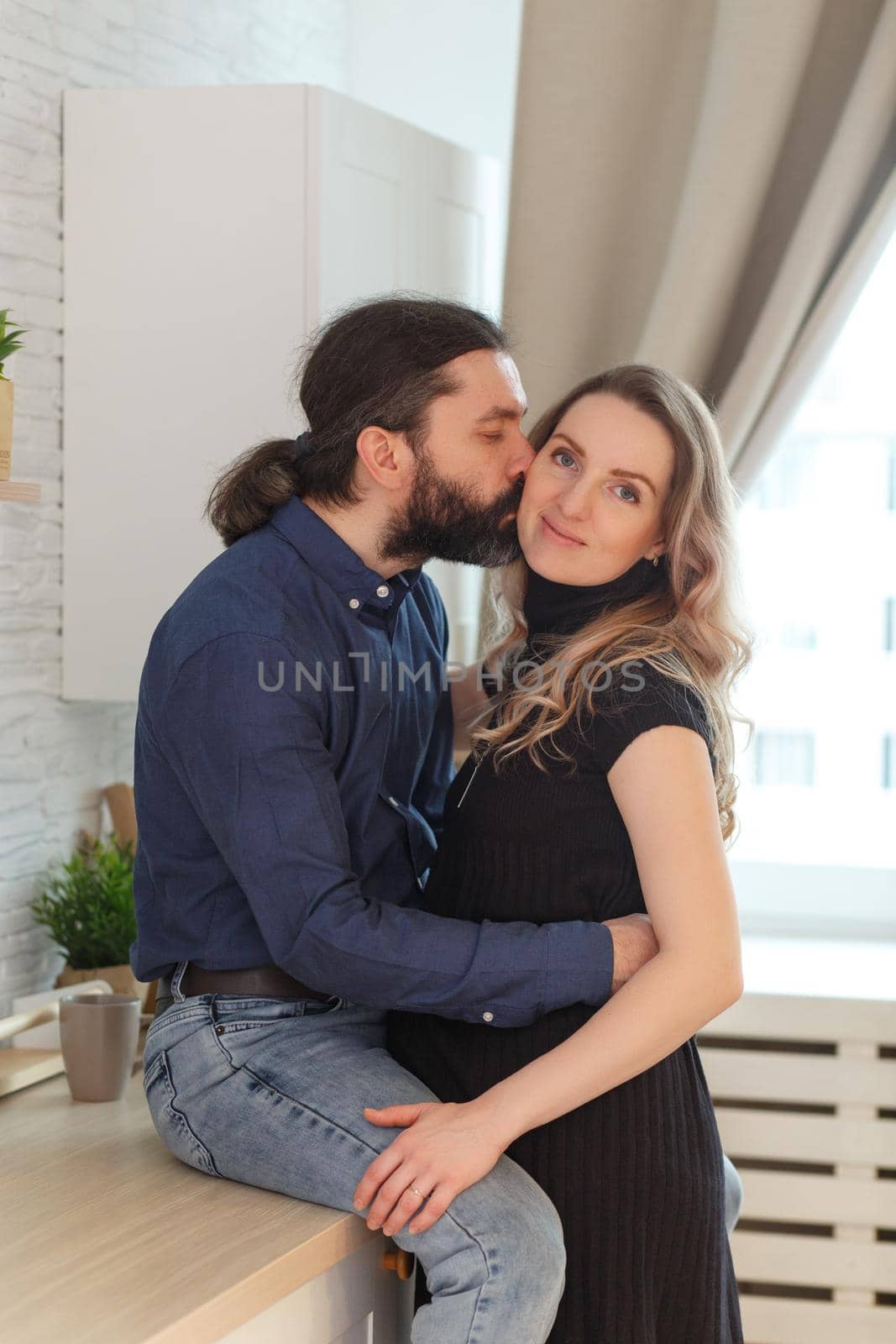 Man embracing pregnant partner in kitchen. Adult family pregnancy concept. Future parents in home outfit embrace standing in the kitchen, looking at each other, kissing. Healthy Lifestyle.