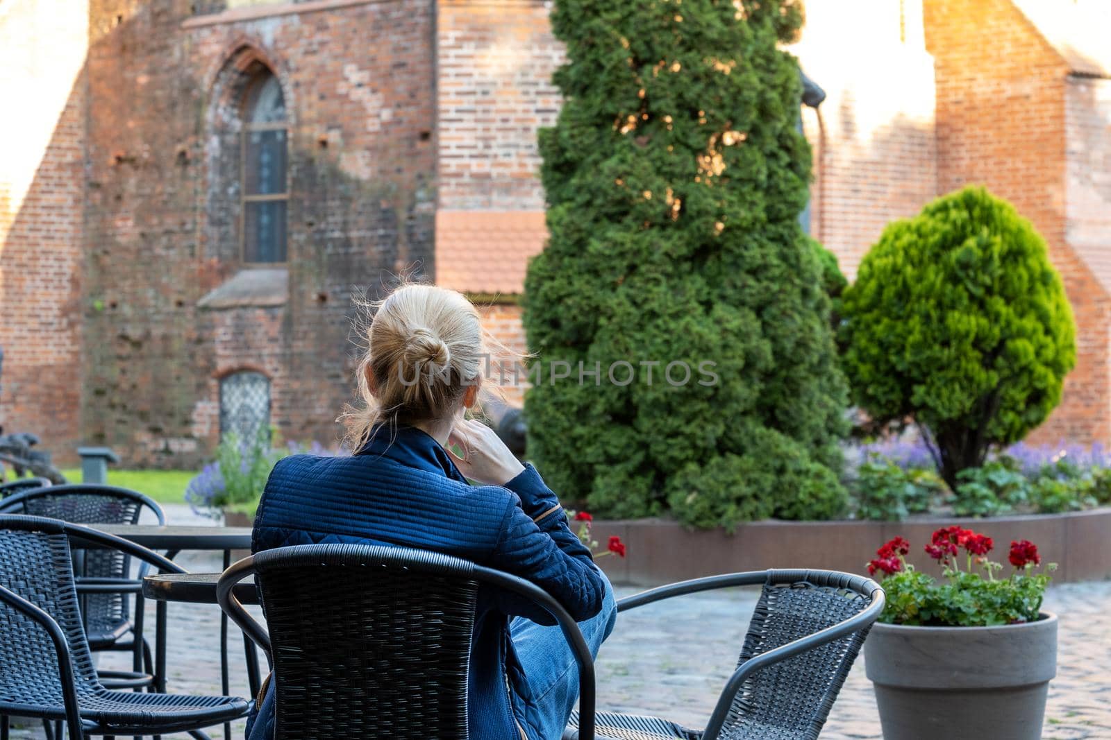 Young blonde woman sits on wicker chair in street cafe and looks at historic red brick building. Selective focus. View from the back.