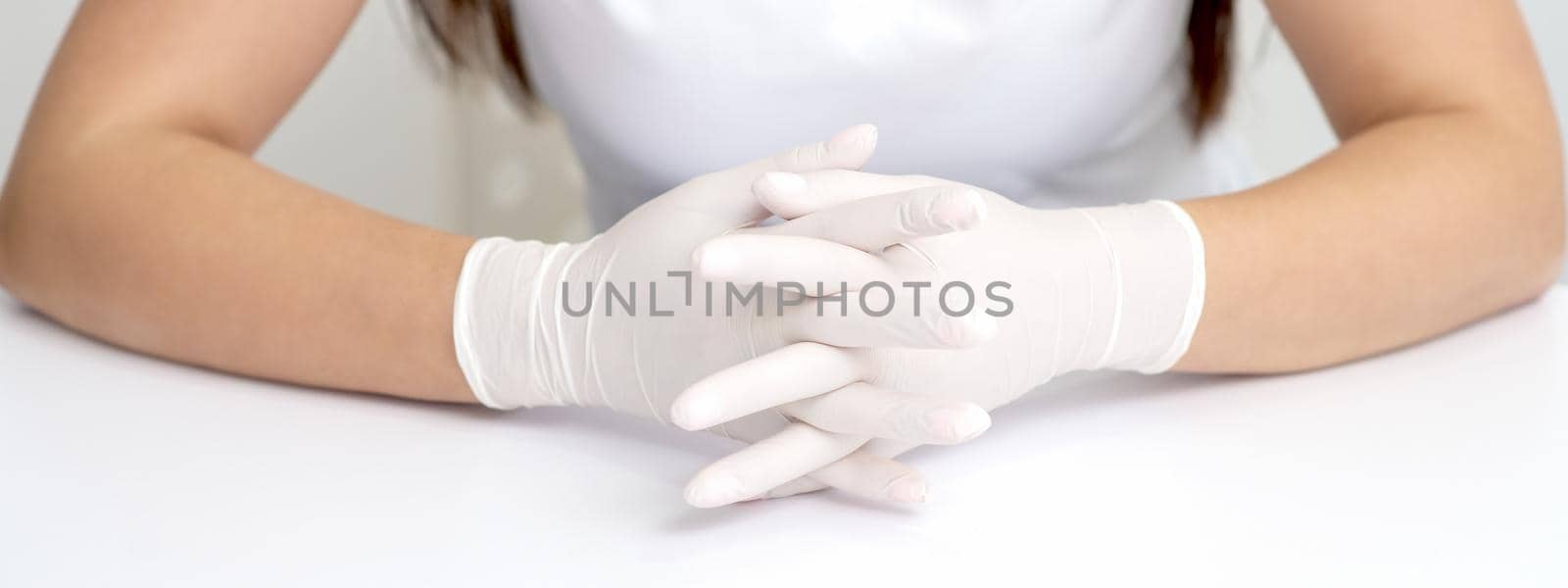 Hands in protective white medical gloves of the woman sitting at the white table. Crossed fingers in gloves