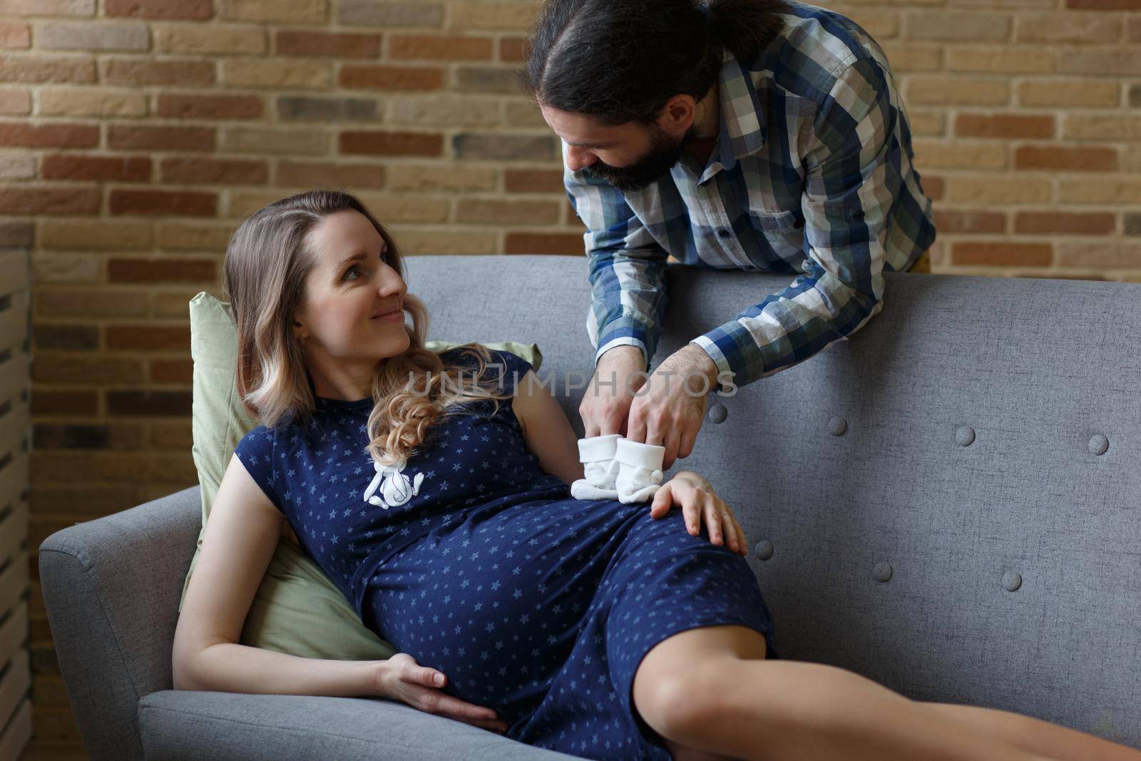 A man and his pregnant wife pose on a sofa in their home. They are holding baby booties that will be worn by their future child. The concept of waiting for a child.