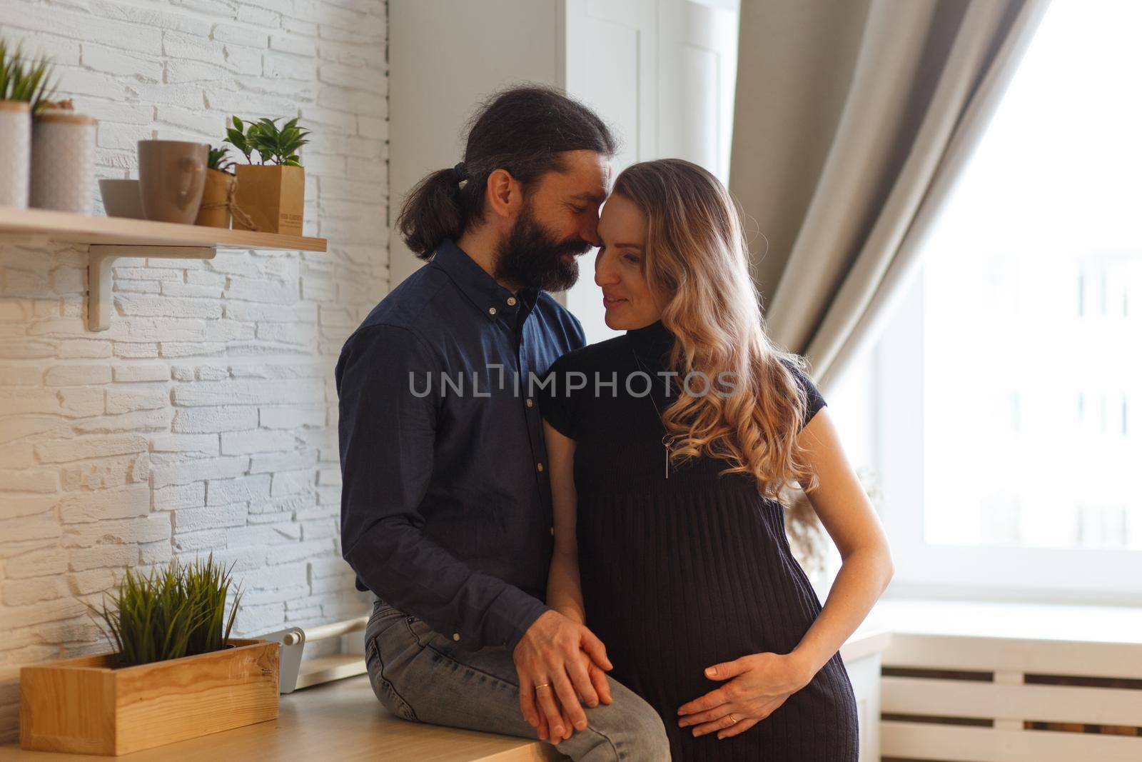 Man embracing pregnant partner in kitchen. Adult family pregnancy concept. Future parents in home outfit embrace standing in the kitchen, looking at each other, kissing. Healthy Lifestyle.