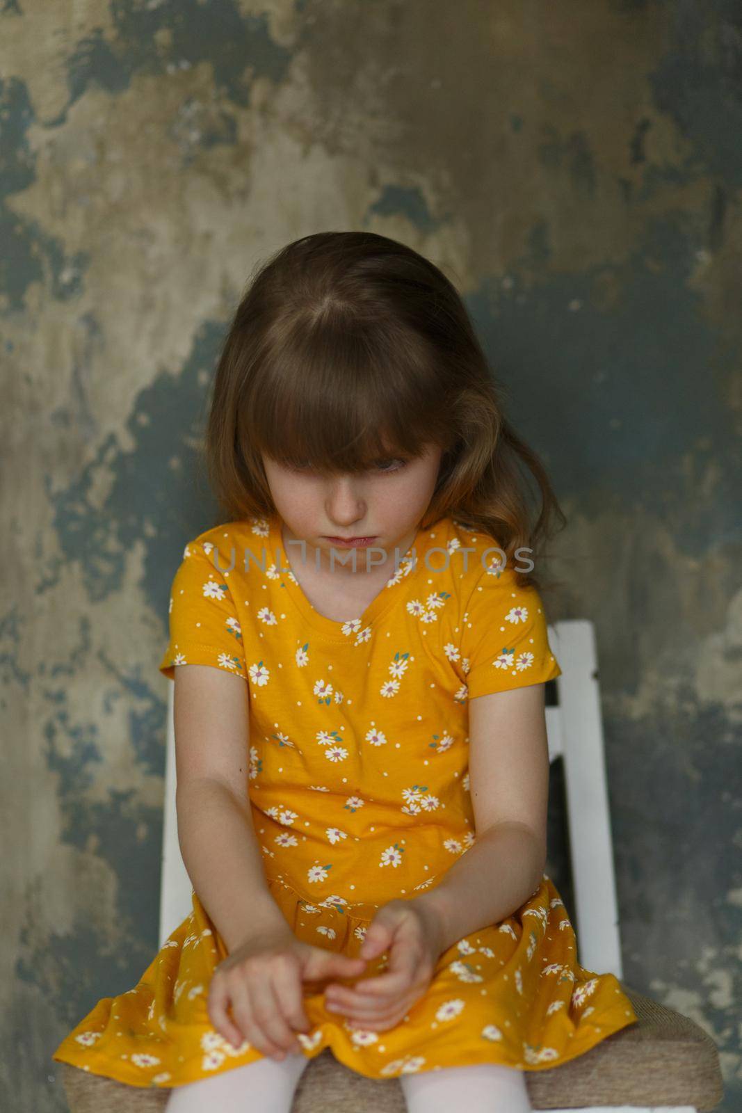 Portrait of a little upset girl in a yellow dress sitting on a chair on an abstract blue background.