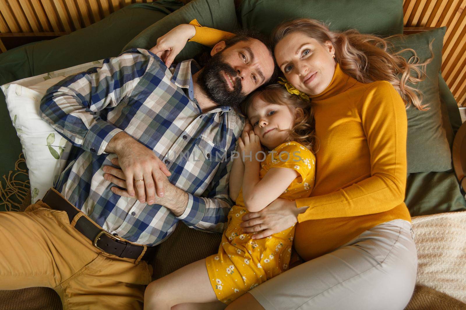 Happy family is having fun in bedroom. Enjoying being together. Parents are tickling their little daughter while lying in bed. Father, daughter and pregnant mother.