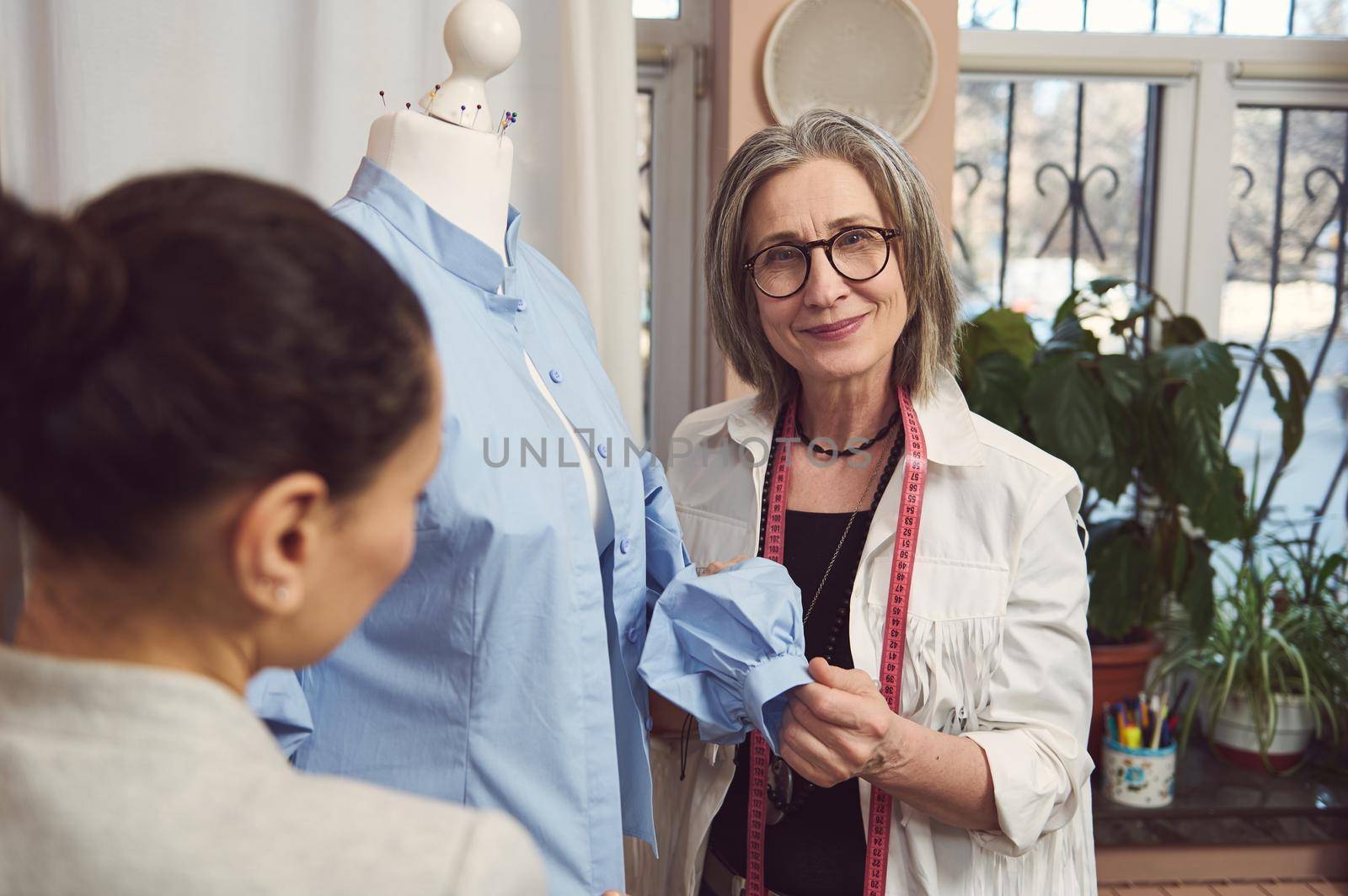 Skilled senior fashion designer takes measurement and works on improvement details of a blue shirt from new collection on a mannequin dummy in a fashion design and tailoring workshop