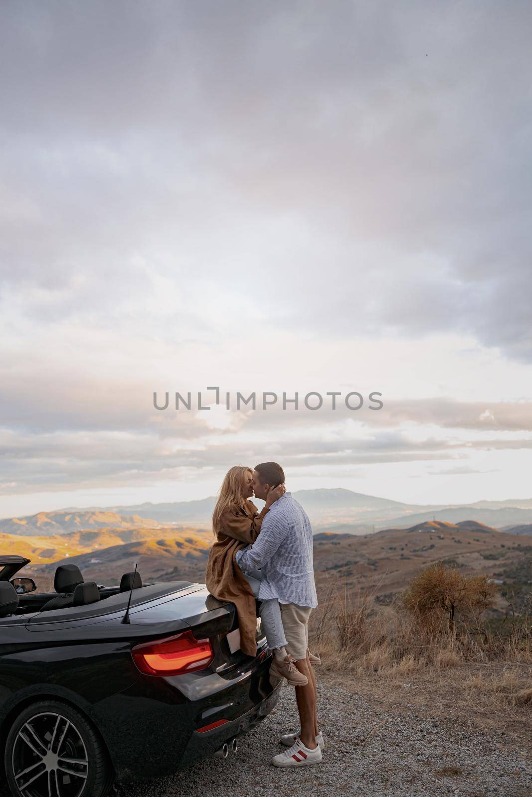 Side view of husband kissing his wife traveling together standing buy cabriolet. Vertical shot