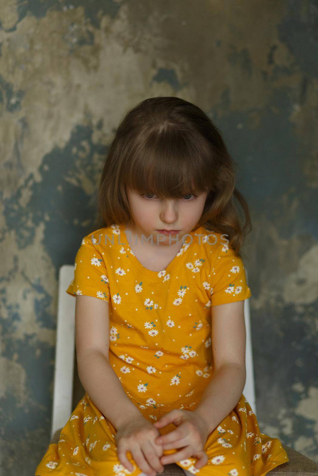 Portrait of a little upset girl in a yellow dress sitting on a chair on an abstract blue background.