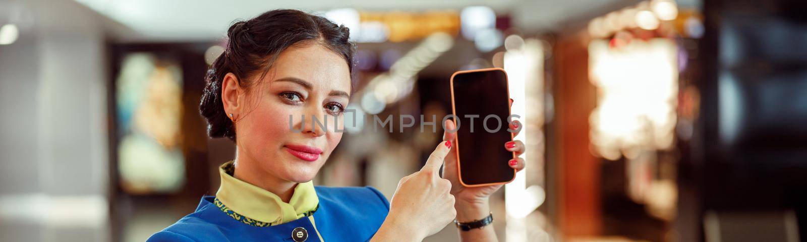 Woman stewardess in aviation air hostess uniform holding mobile phone and smiling while standing in airport terminal