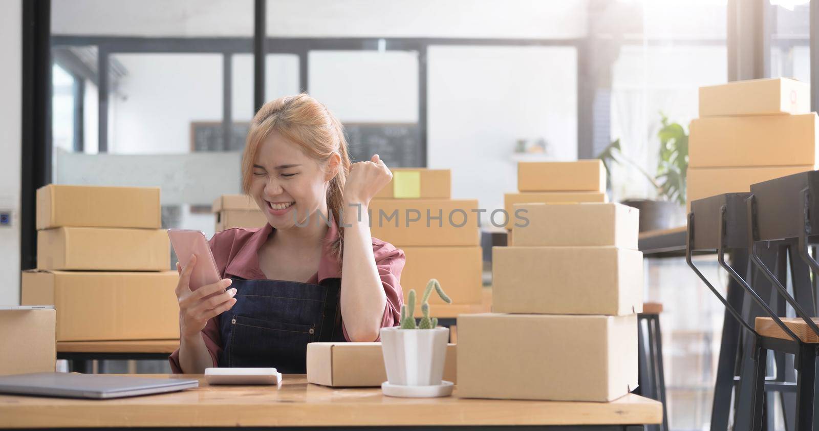 Happy young Asian woman entrepreneur, Smile for sales success after checking order from online shopping store in a smartphone at home office, Concept of merchant business online and eCommerce.