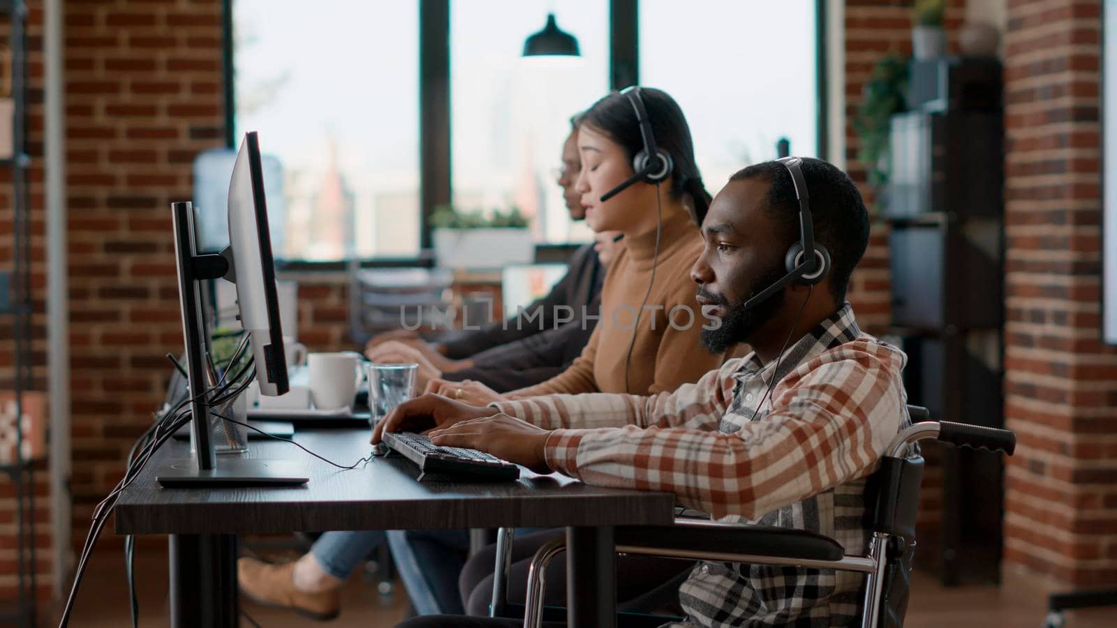 African american employee using headphones to talk to clients, helping with sales and giving telemarketing assistance at call center. Male operatortalking to people in disability friendly office.