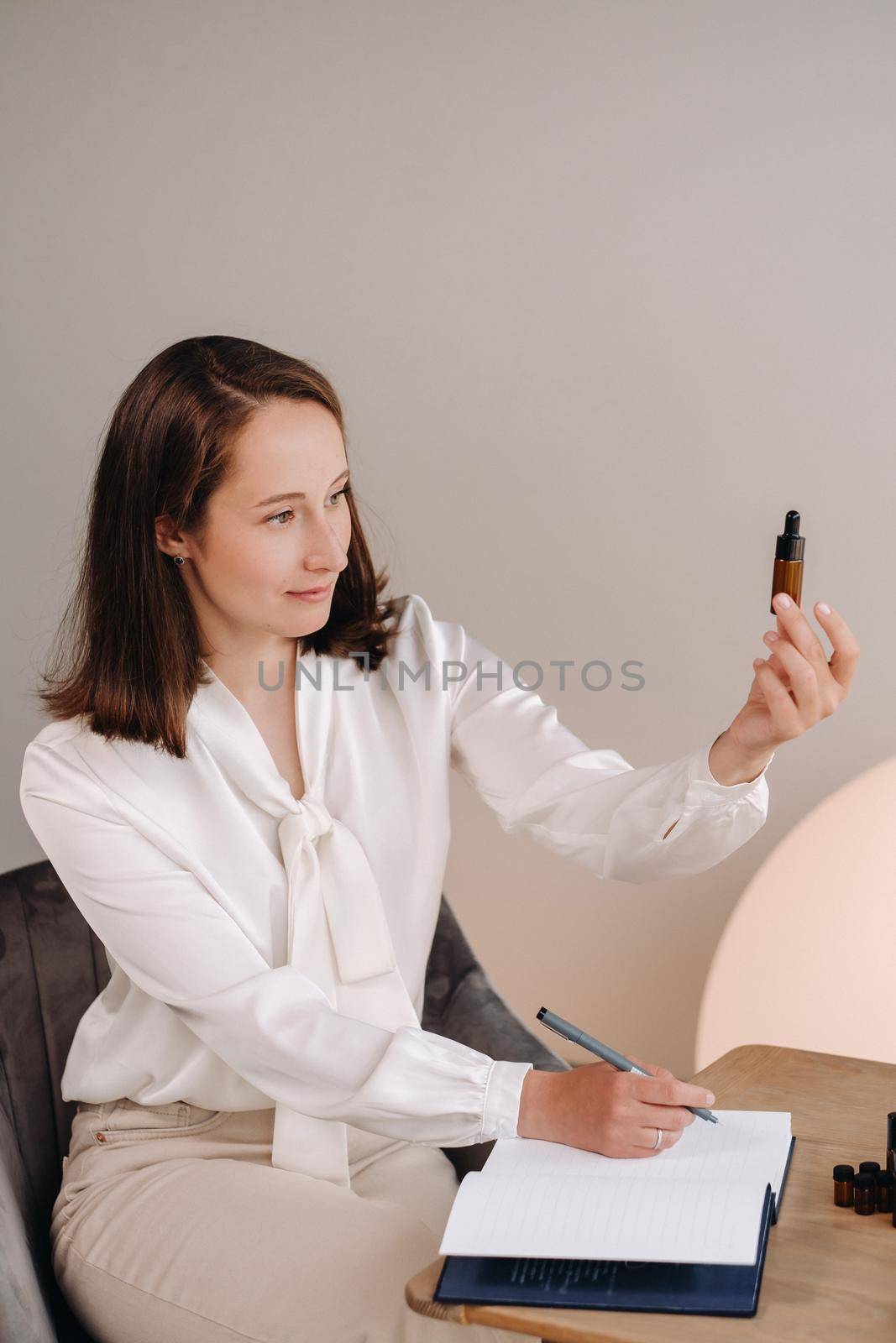 The aromatherapist girl is sitting in her office and holding a bottle of aromatic oil in her hands and writing something down. there are essential oils on the table.