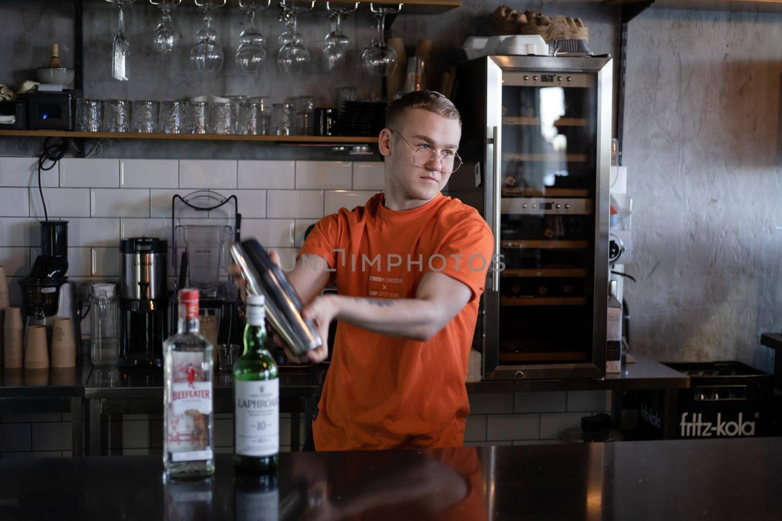 stylish young man hipster in orange t-shirt making mixing a cocktail in a dark loft cafe. alcohol drink in modern bar.